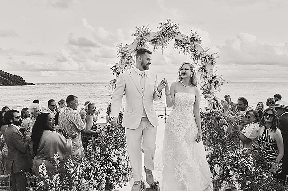 Wedding recessional as bride and groom walk the aisle past cheering guests, bouquet raised, under a floral arch with ocean coastline backdrop