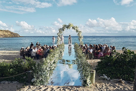 Beach wedding ceremony with a white floral arch and greenery framing a mirrored aisle runner on sand, set against ocean and rocky coastline