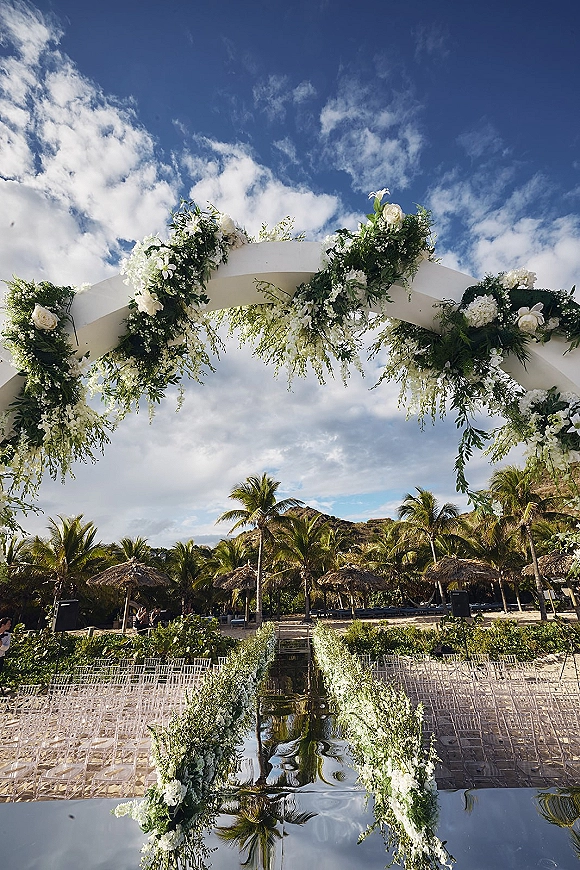 Ceremony aisle design with a mirrored wedding aisle leading to a white floral arch with greenery, set on a beach with palms and mountains