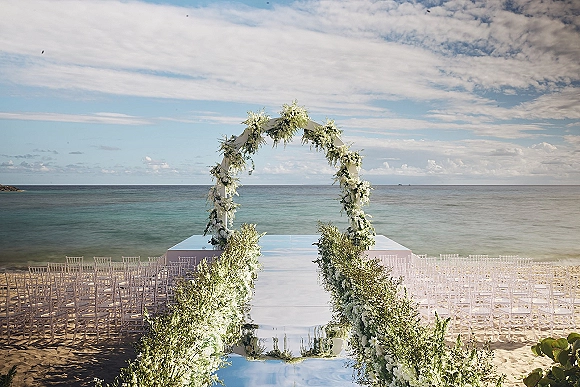 Beach ceremony setup with a round white floral arch, mirrored aisle and clear chairs facing a white stage, set against the ocean horizon