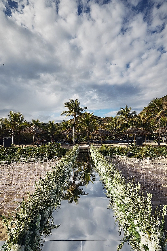 Ceremony aisle design with a mirrored wedding aisle runner reflecting white floral garlands and greenery, set on a sandy beach under palms