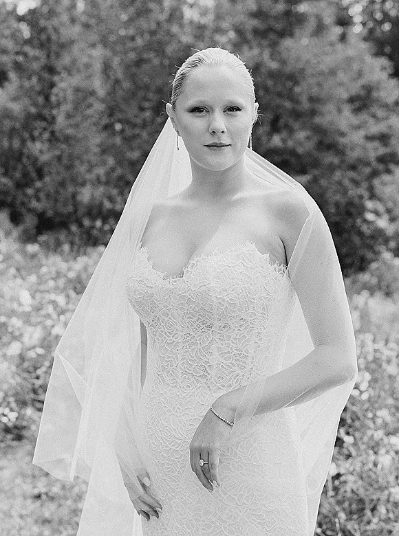 Bridal portrait in black and white of a bride wearing veil over a strapless lace dress, with drop earrings, in a garden setting