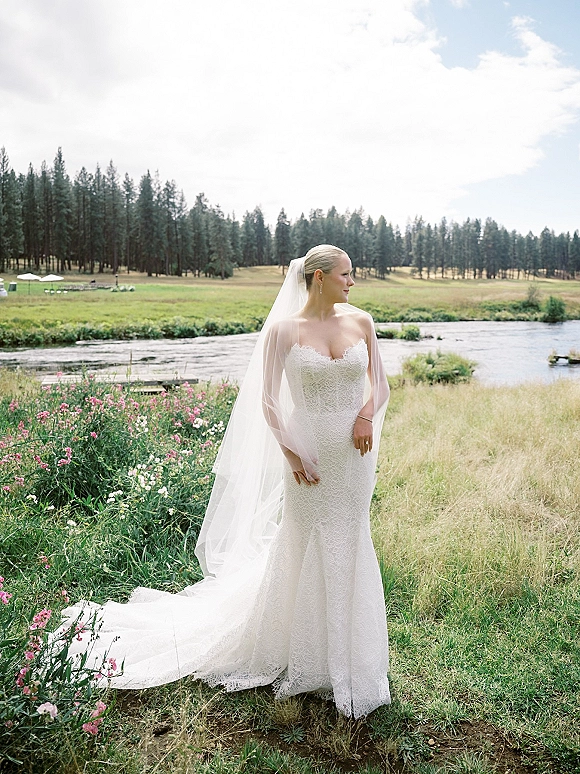 Bridal portrait of a bride in a strapless lace wedding dress with long veil, drop earrings, and ring by a river meadow under clouds