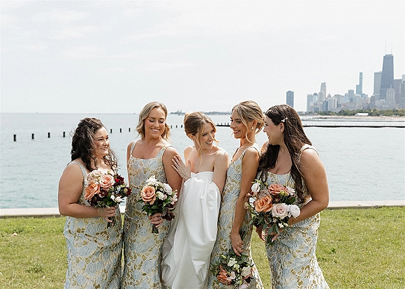 Bridesmaid group photo with bride and bridesmaids laughing, holding bouquets by a lake shoreline with grass lawn and city skyline behind