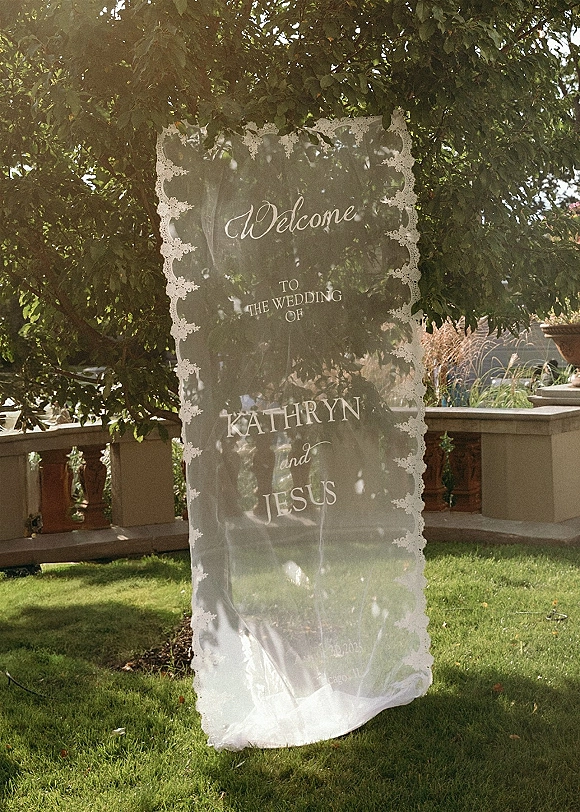 Wedding welcome sign with sheer fabric and calligraphy lettering, trimmed in lace, hanging on a sunlit garden terrace by a stone balustrade