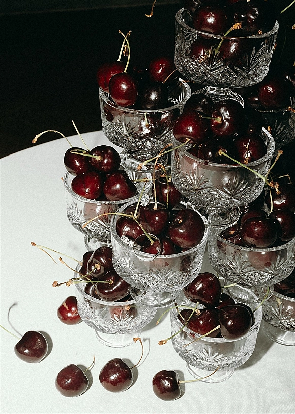 Wedding food styling featuring a cherry dessert bar with cut glass dessert cups and pedestal bowls on a white surface against dark backdrop