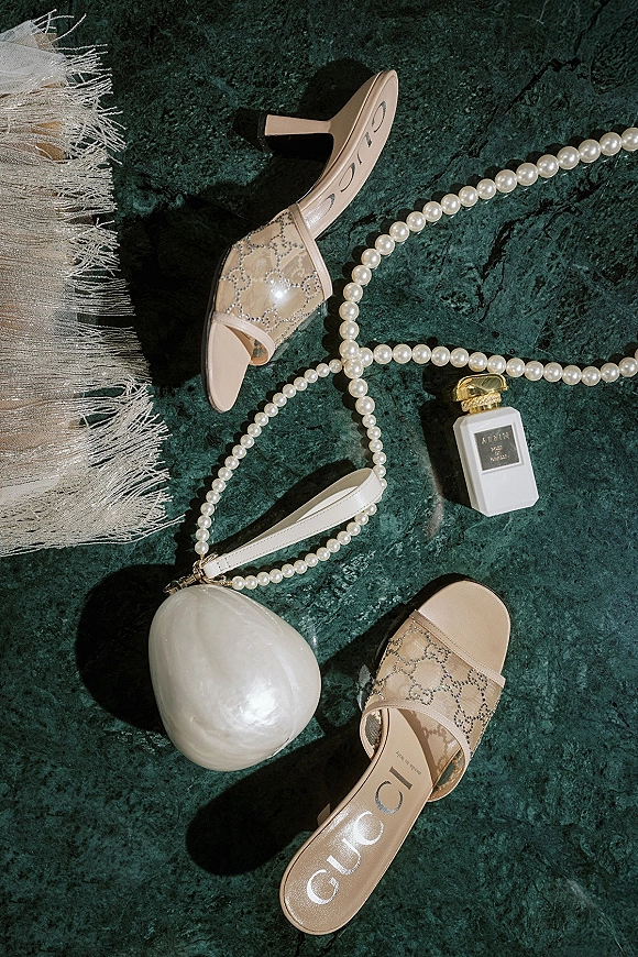 Bridal accessories flatlay with ivory heeled sandals, pearl necklace, perfume bottle and pearl clutch arranged on green marble surface