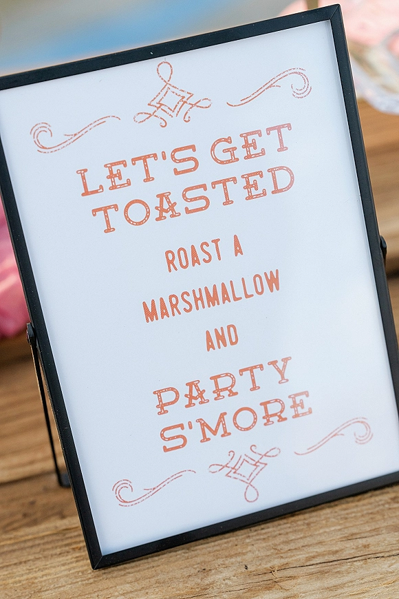 Wedding signage s'mores bar sign in a framed typography print on a tabletop easel, set on a wood table with blurred pink fabric behind