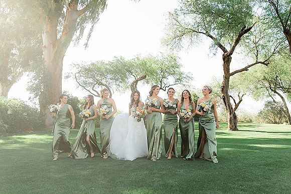 Bridesmaid group photo with bride and bridesmaids in sage green bridesmaid dresses holding bouquets on a green lawn under trees in daylight sky