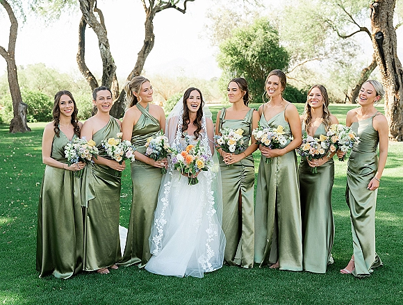 Bridesmaid group photo with bride and bridesmaids in sage green satin dresses holding bouquets on a garden lawn with trees behind