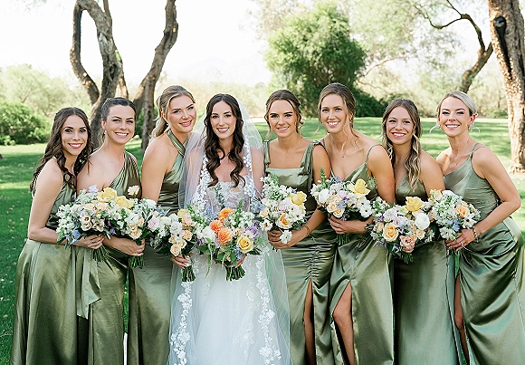 Bridesmaid group photo of bride with bridesmaids in sage green dresses, holding pastel bouquets on a park lawn with trees behind