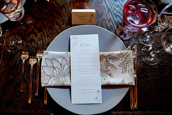 Wedding place setting with wedding menu card on plate, botanical printed napkin accent, mixed flatware, and glasses on a wood table