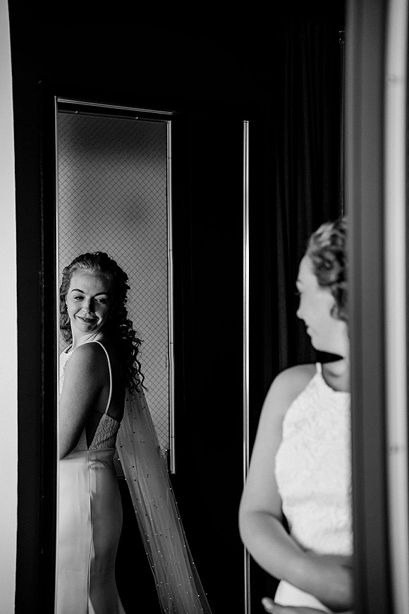 Bridal portrait of a bride looking in mirror, showing a long veil, spaghetti-strap gown, and drop earrings in a dark doorway setting