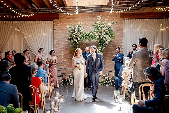 Ceremony recessional as bride and groom walking aisle past candle-lined lanterns, string lights, and a floral arch against brick wall indoors