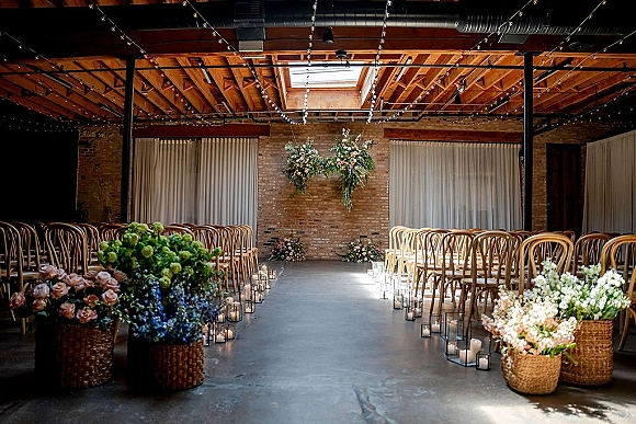 Ceremony setup with an industrial wedding ceremony aisle lined with glass candle holders, wood chairs, hanging florals, and a brick wall backdrop.
