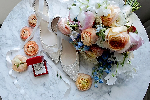 Bridal details flatlay with lace wedding shoes, bouquet, red ring box and wedding rings, earrings and ribbon on a marble table