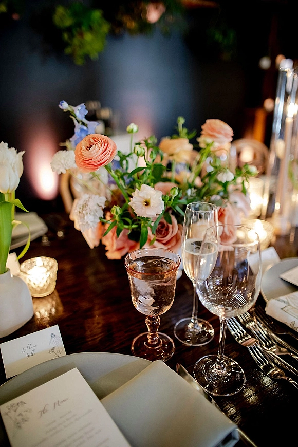 Reception tablescape with wedding table centerpiece of pastel roses and ranunculus, taper candles and glassware on a dark wood table in dim light
