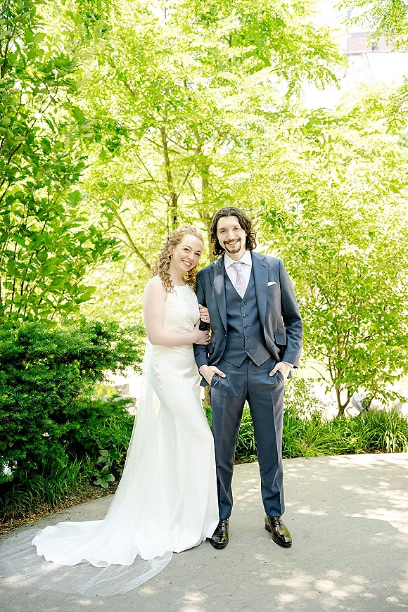 Couple portrait of bride holding groom’s arm in a fitted gown and chapel veil beside him in a three-piece suit on a garden path