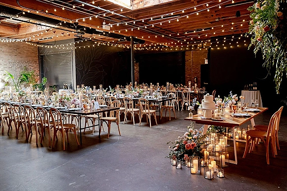 Reception tablescape with long banquet tables wedding under string lights, candles and floral centerpieces, against an industrial brick wall