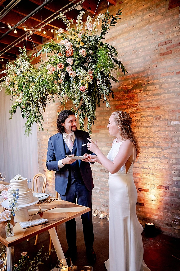 Wedding cake cutting as bride in a minimalist gown and groom in a blue suit slice cake under floral greenery, string lights, brick wall backdrop