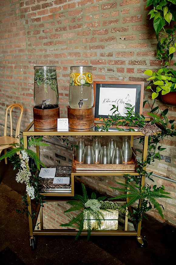 Wedding drink station with glass dispensers of citrus and herb infused water on a gold bar cart, framed sign, greenery, brick wall backdrop