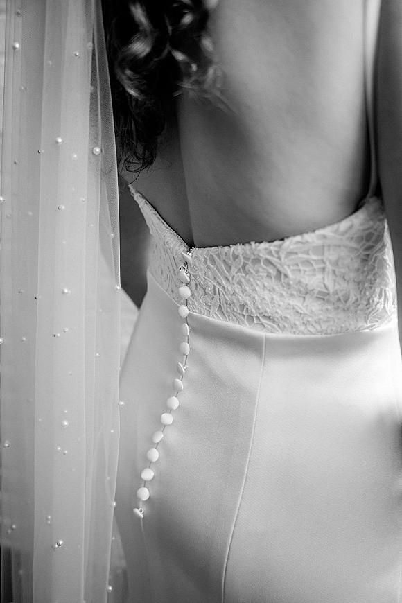 Wedding dress close-up showing lace wedding dress bodice with covered buttons, spaghetti straps, and a pearl veil in soft indoor light