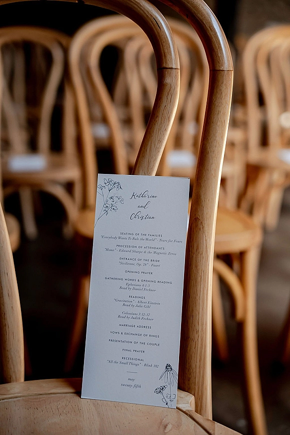 Wedding ceremony program resting on a wooden chair, featuring a black-and-white floral line art design in an indoor seating area