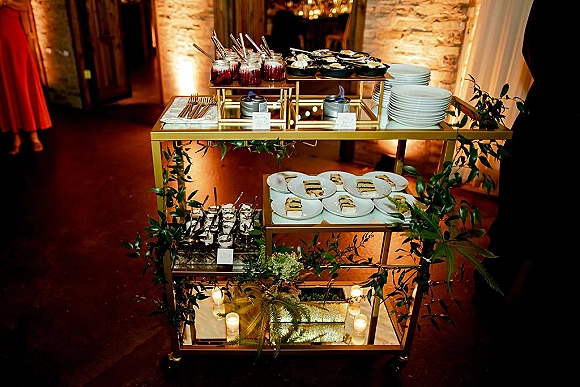 Dessert display on a gold dessert cart with mini parfait cups, candles, and greenery garland set against a brick wall indoors