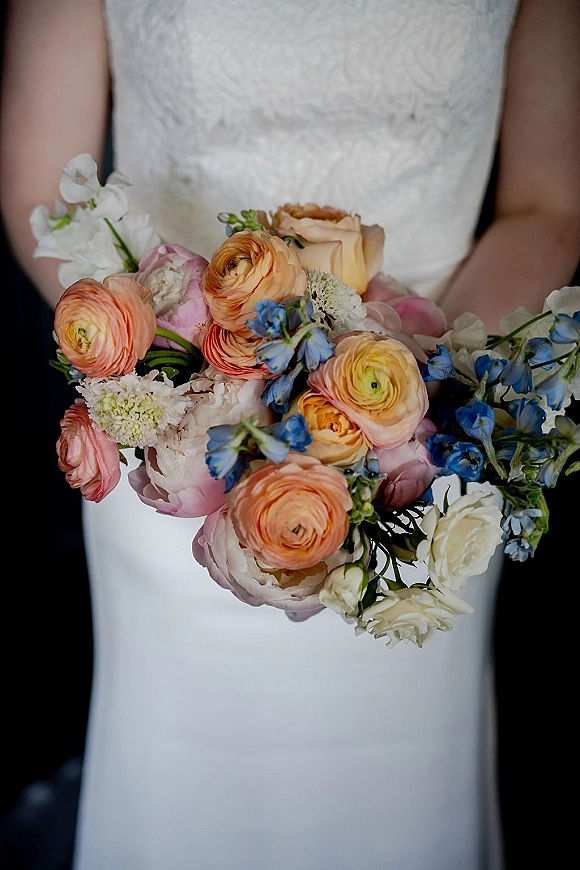 Bridal bouquet of pastel ranunculus with peonies and delphinium, held against a lace wedding dress on a dark backdrop close-up