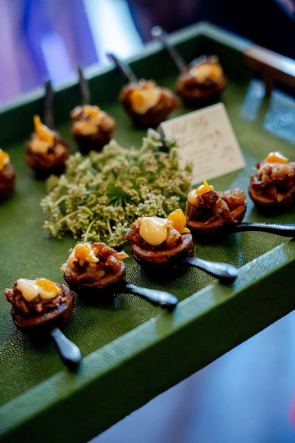 Wedding appetizers and passed hors d'oeuvres arranged as bite-sized canapés and tasting spoons on a green tray with greenery in soft reception light