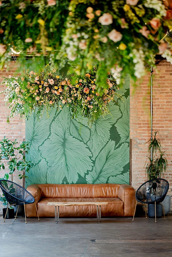 Wedding lounge seating with a wedding lounge area vibe, featuring a brown leather sofa and wire chairs under hanging greenery by a brick wall