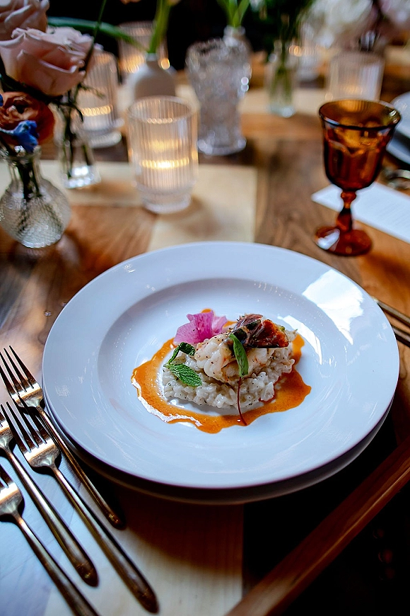 Wedding reception place setting with a plated wedding dinner on a white plate, silver flatware, amber goblet, and candlelit bud vases on wood table