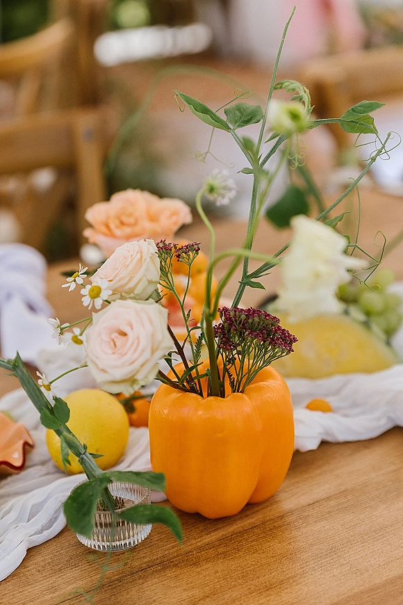 Reception centerpiece with a pumpkin flower centerpiece of roses, wildflowers, and greenery vines, plus citrus fruit on a wood table runner