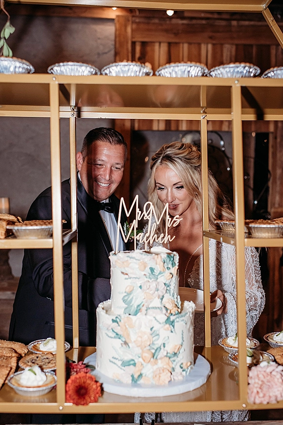 Cake cutting with bride and groom slicing a two-tier floral painted wedding cake on a dessert cart under warm string lights by a wood wall