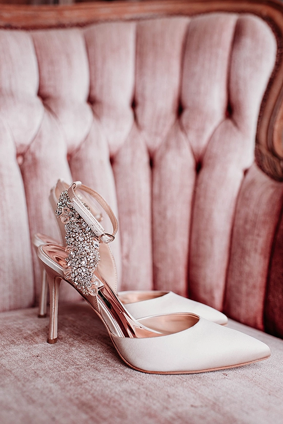Wedding shoes and bridal heels with rhinestone embellishments and ankle straps resting on a blush velvet tufted chair