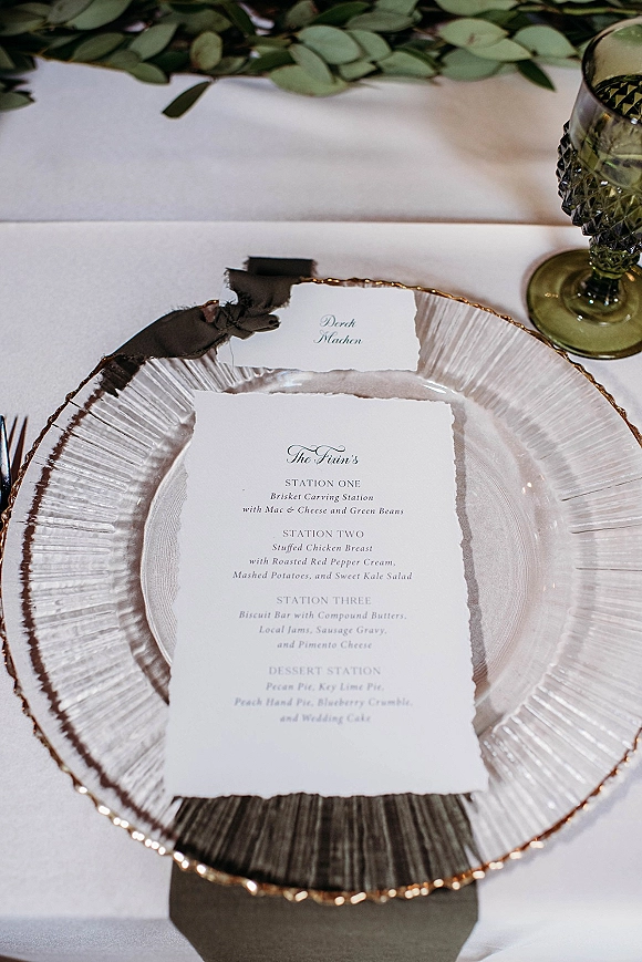 Wedding menu card with deckled edges on a clear glass charger, tied with silk ribbon beside a green goblet on a white tablecloth