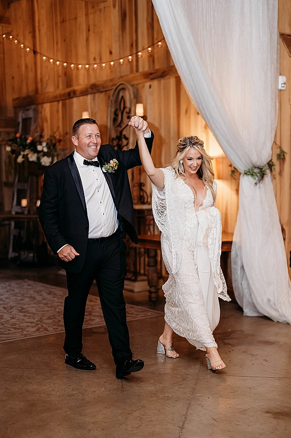 Reception entrance as bride and groom stride in with raised hands under draped fabric and string lights in a rustic barn interior