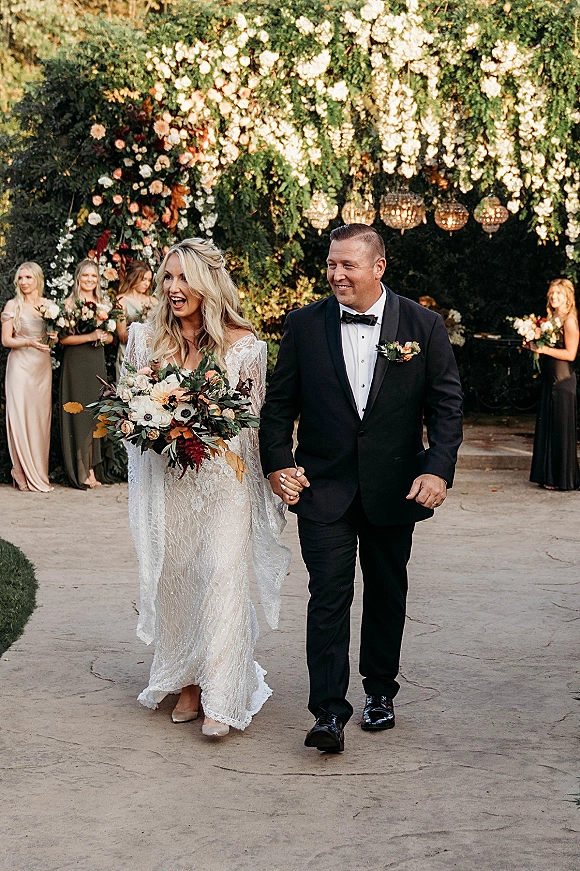 Wedding recessional as bride and groom walk aisle holding hands, her bouquet in a lace gown, under hanging lanterns by a floral arch in a garden