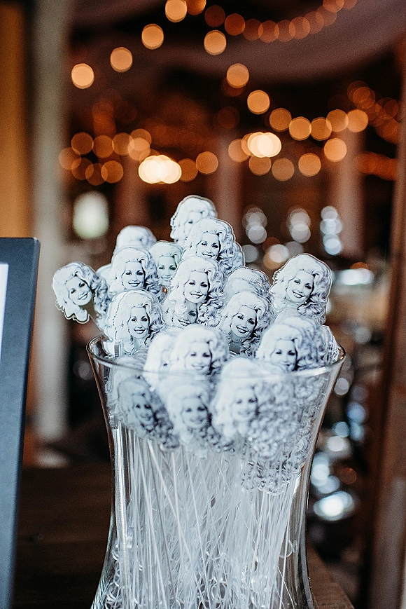 Wedding stir sticks with custom face drink stirrers displayed in a clear glass vase on a bar counter, with soft string light bokeh behind