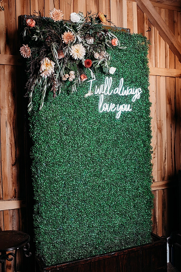 Wedding photo backdrop with boxwood photo backdrop hedge wall, neon sign and peach blush florals framed against a rustic barn wall