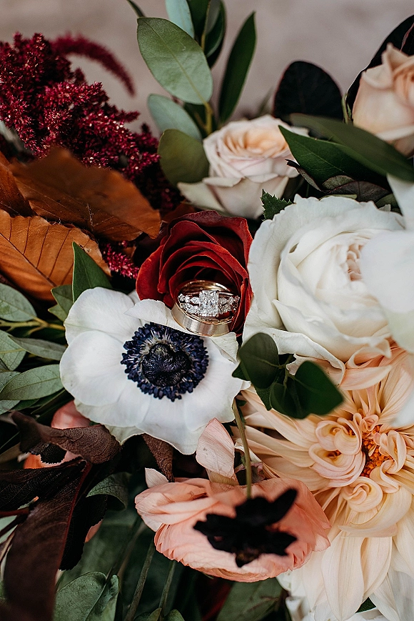 Wedding rings with a diamond engagement ring nestled in a rose and dahlia bouquet, styled on a neutral surface with greenery leaves