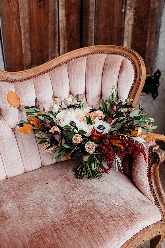 Bridal bouquet with roses, anemones, greenery, and autumn leaves, ribbon-wrapped and set on a vintage velvet chair by a wood plank wall