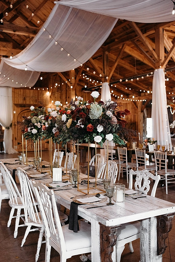 Reception tablescape with elevated floral centerpieces on gold stands, taper candles and glass votives beneath string lights in a wood barn interior