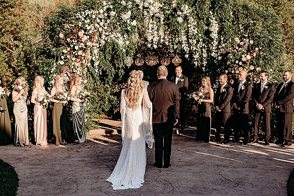 Wedding ceremony moment as bride walks down the aisle toward a floral arch with chandeliers, wedding party lined up in a garden setting