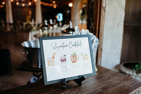 Signature cocktail sign on a stand with framed drink menu and cocktail illustrations on a rustic wood table beneath string lights