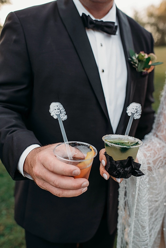 Wedding cocktails held by bride and groom, his and hers cocktails in plastic cups with custom stirrers on an outdoor lawn with trees