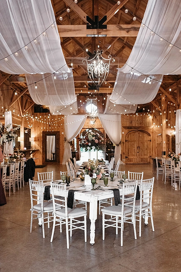 Reception tablescape with barn reception decor featuring white linens, green goblets, candles, and tall florals beneath draped lights in a wood barn