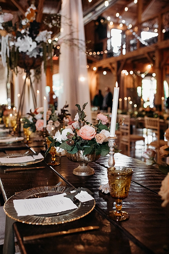 Reception tablescape with wedding head table decor featuring rose centerpiece and taper candles on a dark wood table in a rustic barn with string lights