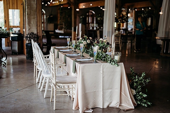 Reception tablescape with farmhouse head table, blush linen, greenery garland, candles and champagne flutes under rustic string lights