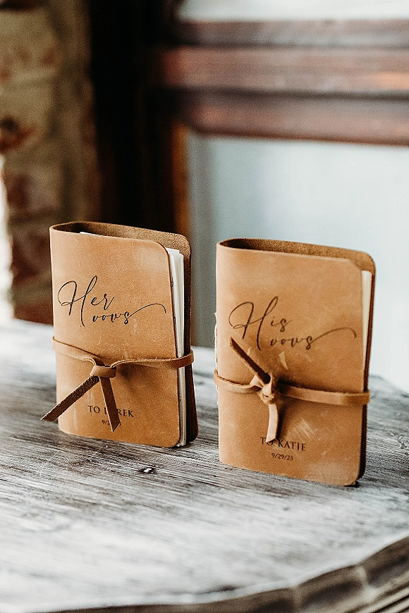 Wedding vows booklets, his and hers vow books in tan leather with wrap ties and calligraphy names and date on a wood tabletop by brick wall window light